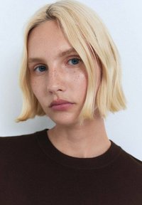 Cheveux blonds courts avec une légère ondulation, de légères taches de rousseur et des yeux bleus clairs. Vêtue d'un pull marron foncé à encolure ronde.