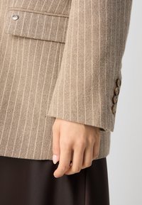 Rękaw beżowej marynarki w drobną prążek z czterema brązowymi guzikami, klapkową kieszenią oraz dłonią spoczywającą blisko brzegu na ciemnej spódnicy.