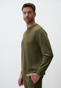 Olijfgroen trui met lange mouwen en een ronde hals, met ribgebreide manchetten en een gladde textuur, gemodelleerd door een man die tegen een neutrale achtergrond staat.