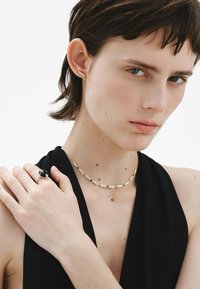 Collar de oro con eslabones texturizados y acentos de piedra negra, combinado con un anillo de oro que presenta una piedra negra redonda, llevado por una persona con cabello corto.