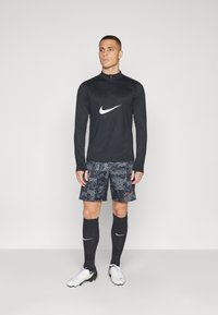 Maglia nera a maniche lunghe Nike con zip, pantaloni corti grigi a fantasia, calze nere alte fino al ginocchio e scarpe da calcio bianche con accenti neri e gialli.