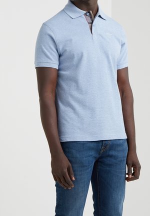 Mężczyzna w jasnoniebieskiej koszulce polo z krótkim rękawem i kołnierzykiem oraz ciemnych niebieskich dżinsach, stojący na jednolitym tle.