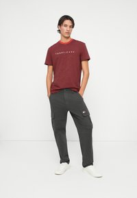 Rød og marineblå stripet t-skjorte med "TOMMY JEANS"-logo, parret med mørkegrå cargo-bukser. Hvite sneakers fullfører antrekket.