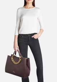 Borsa a tracolla in pelle burgundy con una finitura testurizzata, dotata di una robusta maniglia a catena dorata e di un logo. Il modello indossa una maglietta bianca e jeans scuri.