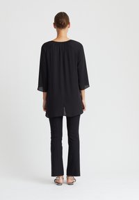 Schwarzes Langarmoberteil mit gerafftem Ausschnitt, kombiniert mit schwarzen ausgestellten Hosen und Sandalen mit offenen Zehen. Glatter Stoff, minimalistisches Design.