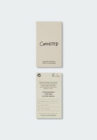 Etiqueta de algodón sostenible en papel reciclado beige, con texto en negro. En el frente dice "Comprometido", y en la parte posterior enumera prácticas ecológicas y detalles de cuidado.