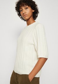 Weißer Ripp-Strickpullover mit kurzen Ärmeln, rundem Ausschnitt und weichem Material. Kombiniert mit olivgrünen Hosen, die eine lockere Passform zeigen.