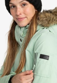 Mint green winter jacket with a fur-lined hood, snap and zip closures, and a black fabric patch on the sleeve labeled "ROXY."