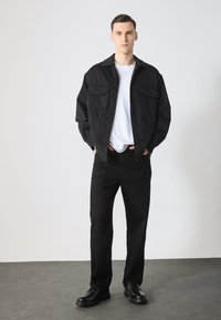 Schwarze langärmelige Hemdjacke mit zwei Brusttaschen, kombiniert mit einem weißen T-Shirt, schwarzen Jeans und schwarzen Schuhen, stehend auf einer grauen Fläche.