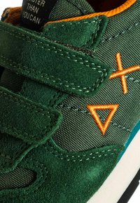 Sneaker verde con strap in velcro, simboli ricamati arancioni e profilo arancione lungo l'apertura della scarpa.