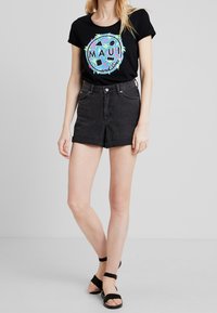 Camiseta negra con un gráfico colorido de "MAUI", combinada con pantalones cortos de mezclilla negros de cintura alta. El calzado consiste en sandalias negras con tiras al tobillo.