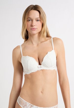Reggiseno in pizzo bianco con coppe imbottite, bordi smerlati e un piccolo fiocco al centro. Spalline regolabili e tessuto con trama.