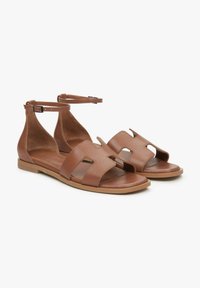 Bruine leren sandalen met twee brede banden met uitsnijdingen, enkelbandjes met een zilveren gesp en een vlakke tankleurige zool.