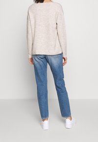 Beige gestrickter Pullover mit lockerer Passform und abgesenkten Schultern, kombiniert mit hellblauen geraden Jeans und weißen Sneakers.