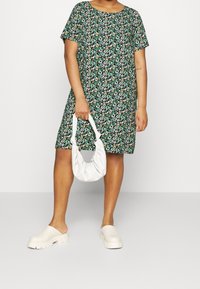 Blommig kortärmad klänning i grönt med flerfärgade blommor, kombinerad med en vit, mjukviktig handväska och kraftiga krämfärgade träskor.