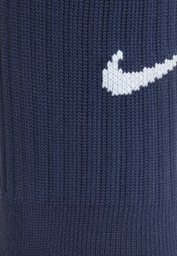 Námořnickomodrá pletená ponožka s bílým logem Nike swoosh. Textura je žebrovaná, což zdůrazňuje její elastický materiál a tvar.