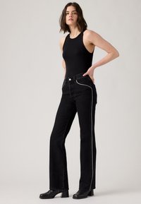 Zwarte geribbelde tanktop met zwarte denim flared jeans. De jeans hebben witte zijnaden en voorzakken. Model draagt zwarte haklaarzen.