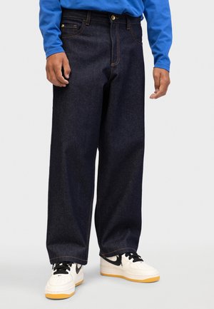 Personne portant un jean large foncé, des baskets blanches et noires avec des semelles en gomme, et une chemise bleue à manches longues, debout sur un fond uni.