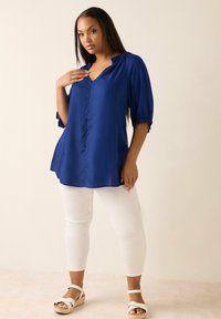 Blå satinblus med krage och knappslå framtill, med trekvartslånga puffärmar. Kombinerad med vita figursydda byxor och beige kilklacksandaler.