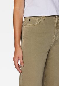 Primo piano della mano di una persona appoggiata accanto a dei jeans a vita alta color beige e al bordo inferiore di una camicia bianca.