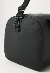 Black nylon bag with a smooth texture, rounded shape, adjustable strap, and a distinctive metal clip on the front for closure.