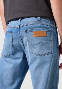 Jeans de mezclilla azul claro con una textura suave, que cuentan con una etiqueta de cuero marrón y un distintivo cosido en los bolsillos traseros.