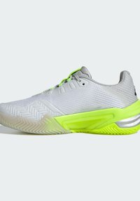Zapatilla deportiva blanca con parte superior de malla, acentos plateados y suela y cordones de un vibrante verde neón; presenta un patrón de banda de rodadura en zigzag en la suela exterior.