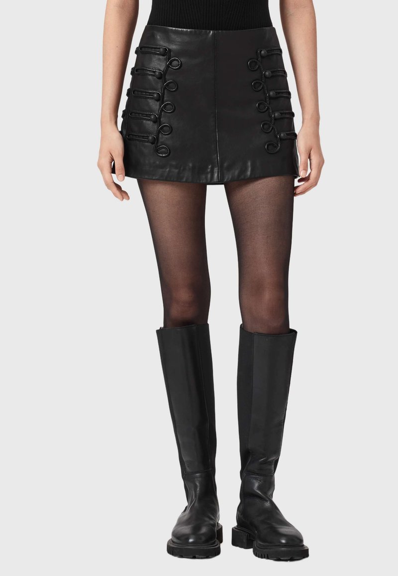 Czarna mini spódnica z skóry z dekoracyjnymi pętlami na przednich panelach, zestawiona z przejrzystymi czarnymi rajstopami i czarnymi kozakami za kolano.