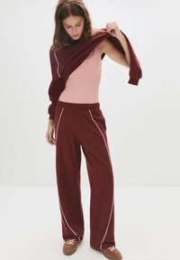 Burgundy træningsbukser med hvide pipedetaljer, parret med en pink top. Tøjet har et afslappet design og en løs pasform.