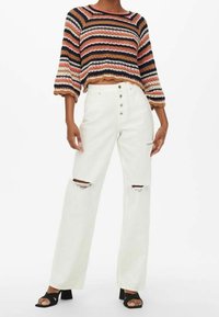 Sweter dzianinowy w paski, krótki, w kolorach czerwonym, czarnym i beżowym; zestawiony z białymi szerokimi spodniami z dziurami i zapięciem na pięć guzików. Czarne szpilki.