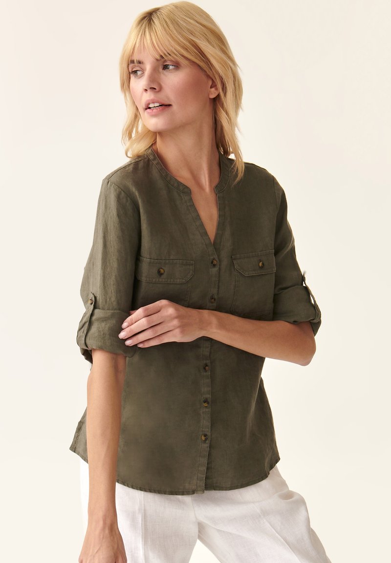 Camicia di lino verde oliva con colletto, apertura con bottoni e due tasche sul petto. Maniche arrotolate. Texture liscia con una vestibilità rilassata.