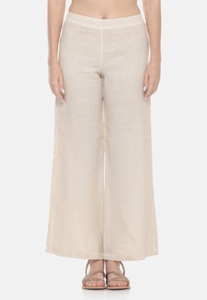 Donna con pantaloni beige in lino a vita alta e gamba larga e sandali beige con cinturini, in piedi con le mani rilassate lungo i fianchi.