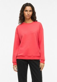 Sweat-shirt corail en tissu jersey, avec un col rond, des épaules tombantes, des manches longues et une coupe décontractée. Porté avec un pantalon noir.