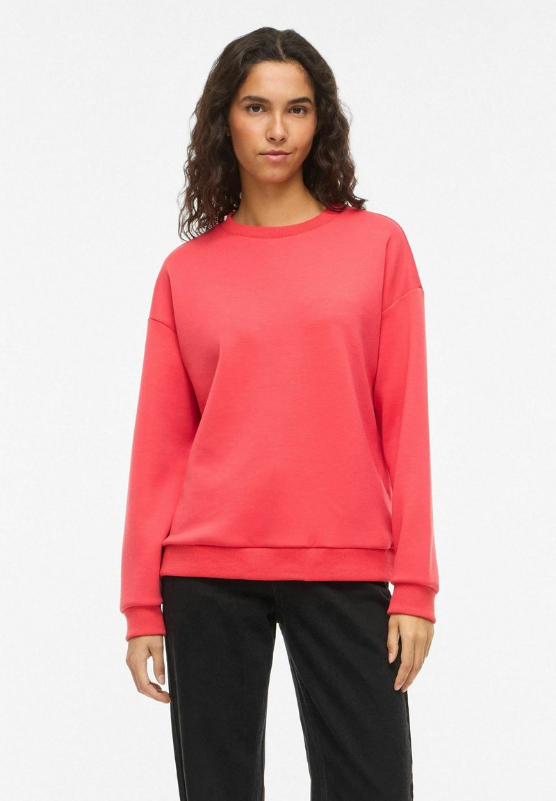 Sweat-shirt corail en tissu jersey, avec un col rond, des épaules tombantes, des manches longues et une coupe décontractée. Porté avec un pantalon noir.