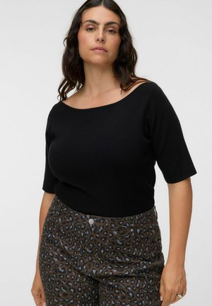 Femme aux longs cheveux foncés portant un haut noir à manches courtes et un pantalon taille haute imprimé léopard marron et noir.