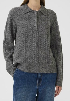 Femme portant un pull gris à torsades avec un col et trois boutons, associé à un jean bleu, debout avec une main dans la poche.