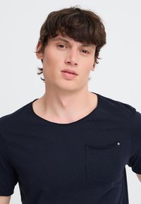 Young man with short brown hair wearing a black t-shirt with a small chest pocket, looking slightly to the side against a white background.