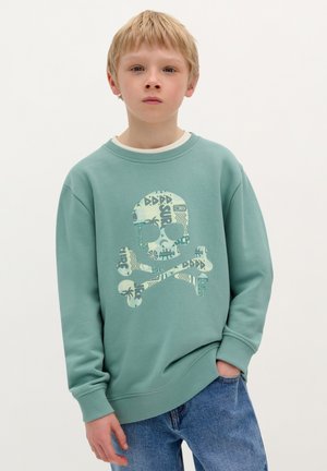 Mintgroene sweatshirt met een groot, meerkleurig doodskop en gekruiste botten graphic. Ronde hals en geribbelde manchetten, gecombineerd met blauwe jeans.