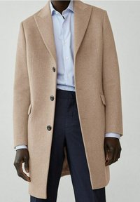 Beige wollen jas met een gepaste uitstraling, met reverskraag en drie zwarte knopen. Draagt over een lichtblauw gestreept shirt en donkere broek.