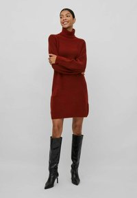 Abito a collo alto lavorato a maglia rosso con maniche lunghe, caratterizzato da spacchi laterali. Abbinato a stivali neri in pelle lucida al ginocchio.