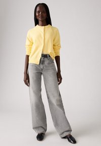 Een gele, knoopsluiting cardi van zachte stof met lange, opgerolde mouwen, gecombineerd met hoog afgegraven, wijde grijze jeans. Zwarte schoenen maken de outfit compleet.