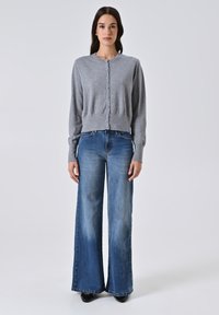 Donna in piedi di fronte, indossa un cardigan grigio chiaro con bottoni, jeans azzurri a gamba larga, scarpe nere e orecchini dorati, su uno sfondo grigio chiaro.