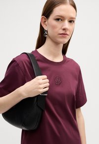 Burgundiško medvilninio marškinėlių su tekstūruotu logotipu, derinami su juodu odiniu rankiniu krepšiu, turinčiu lygią apdailą ir išlenktą formą.