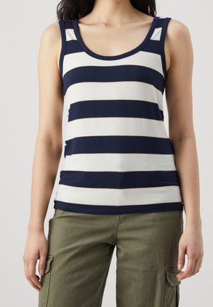 Kvinde iført navy og hvid, vandrette, ærmeløse tanktop og olivengrønne cargobukser, stående foran en ensfarvet baggrund.