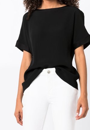 Vrouw draagt een loszittend zwart shirt met korte mouwen, lichtjes ingestopt in witte hoge taillebroek met een zichtbare knoop.