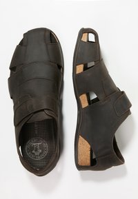 Dark brown leather sandals featuring an open-toe design, adjustable straps, and cork-accented soles with perforated insoles for breathability.