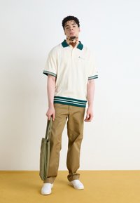 Kremfarget polo skjorte med grønne striper ved kragen og mansjettene, sammen med brune bukser og hvite sko. Holder en grønn totebag.
