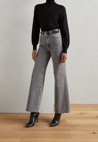 Ein schwarzer Rollkragensweater kombiniert mit grauen weit geschnittenen Jeans und schwarzen, spitz zulaufenden Absatzstiefeln. Texturierte Strick- und Denim-Materialien kommen zum Einsatz.