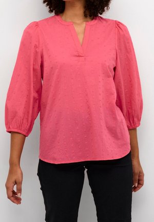 Nainen, joka on pukeutunut korallinpunaisella teksturoidulla puseroon, jossa on kolmen neljänneksen mittaiset puhvimaiset hihat, ja mustiin housuihin, seisoo valkoista taustaa vasten.