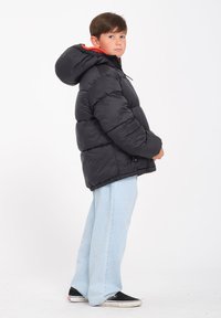 Schwarze Daunenjacke mit Kapuze und orangefarbenem Futter, mit gestepptem Design, lässiger Schnitt, getragen über hellblauen weiten Hosen und schwarzen Schuhen.